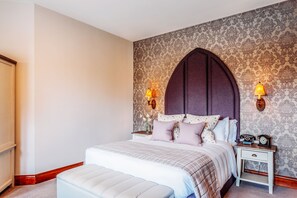 A bedroom with a large bed, a nightstand with a clock, and a wall with patterned wallpaper. - Stanbrook Abbey (Worcester)