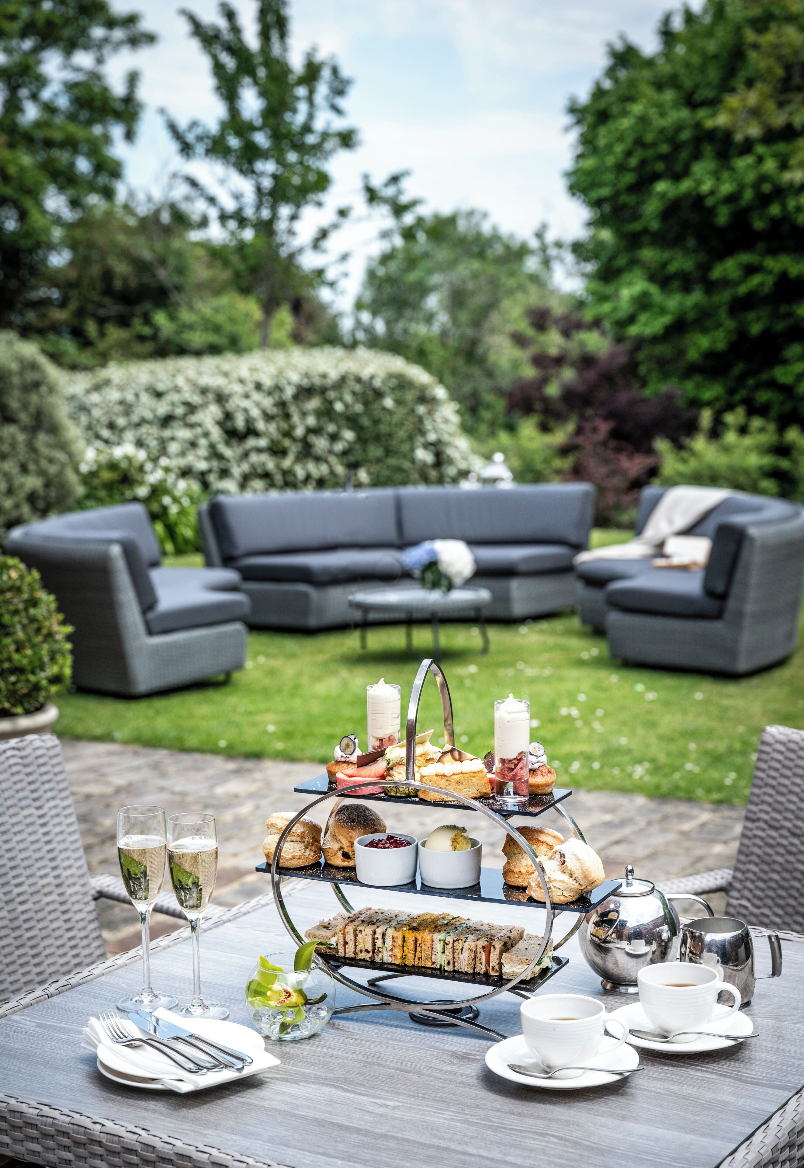 A garden patio with a three-tiered tea service, two glasses of champagne, and a table set for a meal.