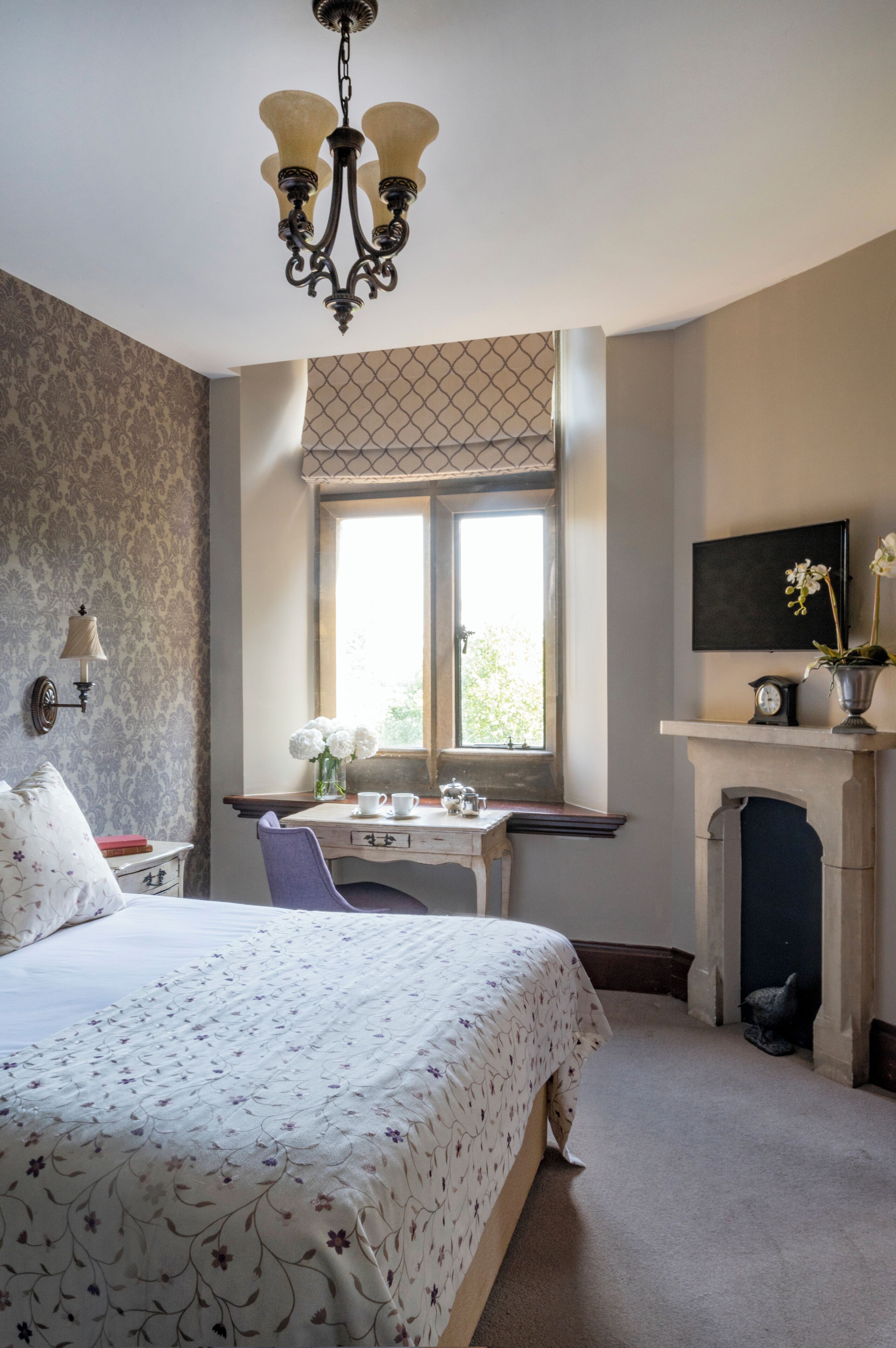A bedroom with a bed, a desk, a window, a fireplace, and a chandelier.