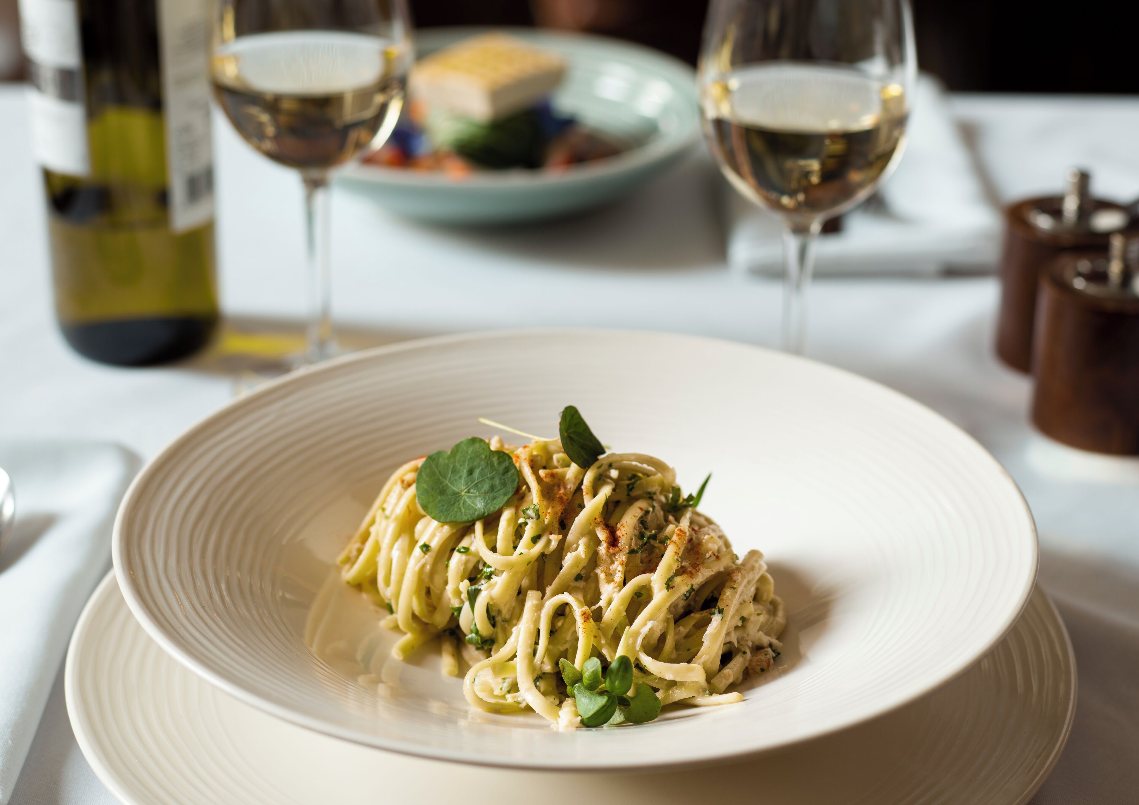 Un plato de pasta con decoración, dos copas de vino blanco y una botella de vino sobre la mesa.