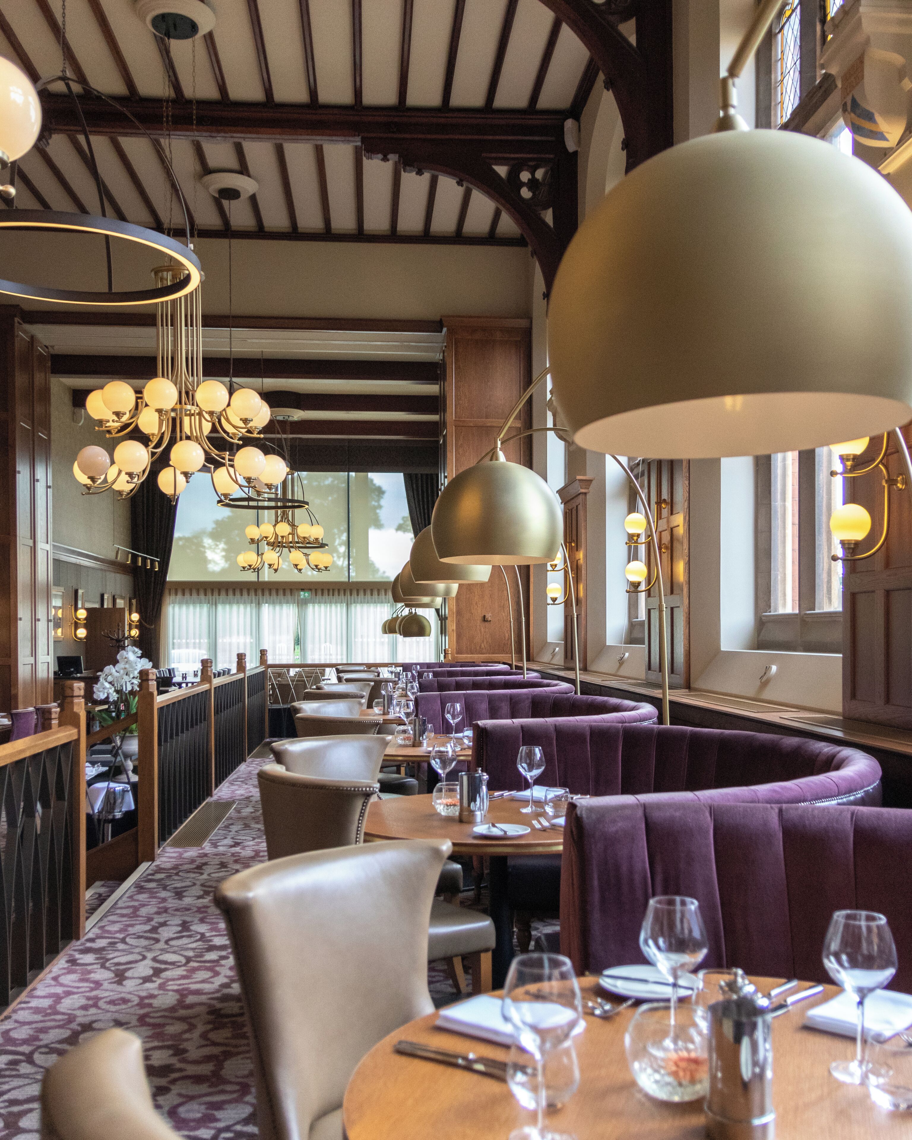 A restaurant with high ceilings, exposed beams, and a variety of pendant lighting fixtures. Tables are set with wine glasses and cutlery, and there are cushioned seating areas.