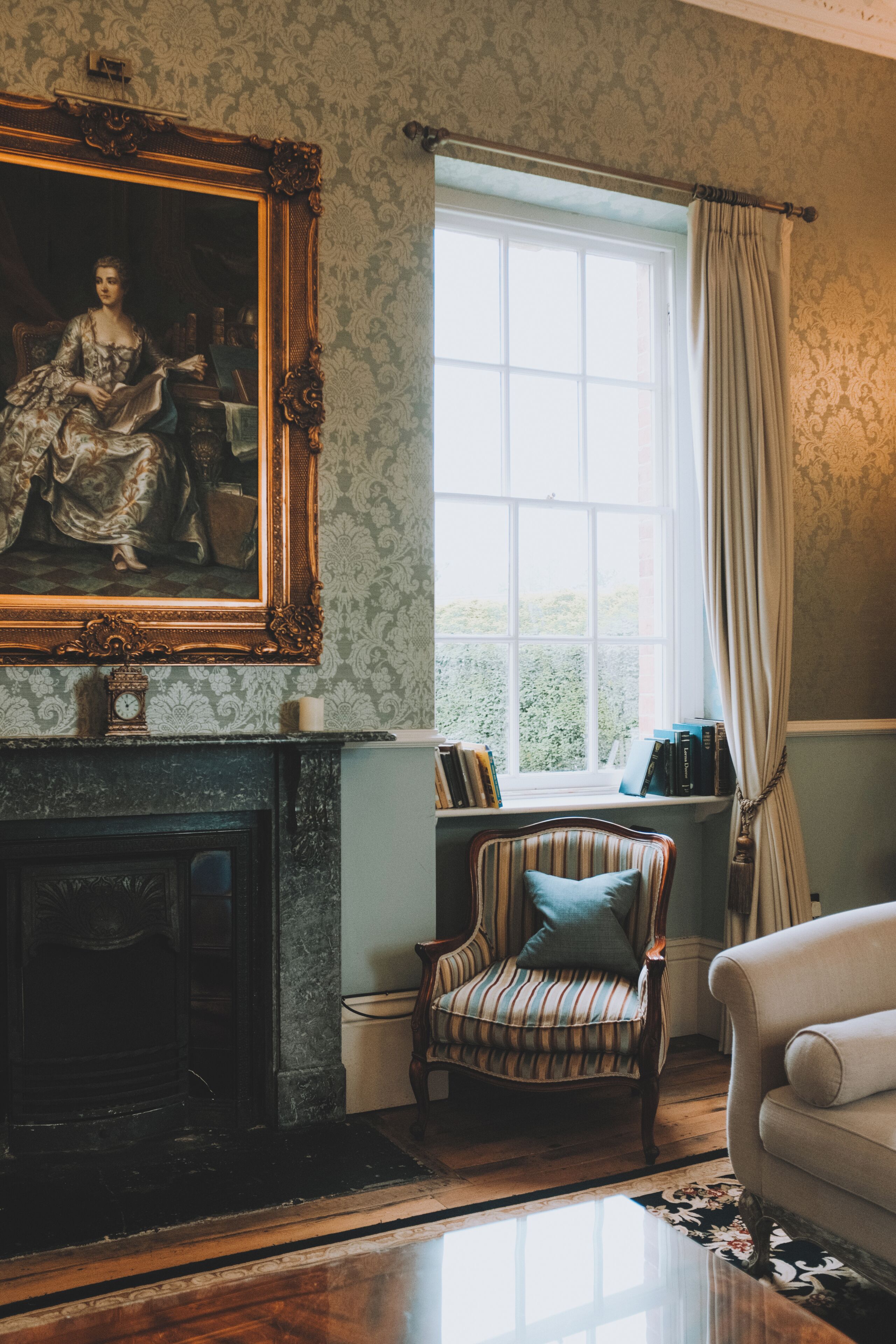 A room with a fireplace, a large framed portrait, a window with curtains, a striped armchair, and a sofa.