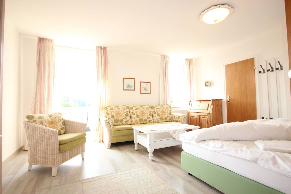A well-lit room with a bed, a green sofa, a wicker armchair, a white coffee table, and a wooden cabinet.