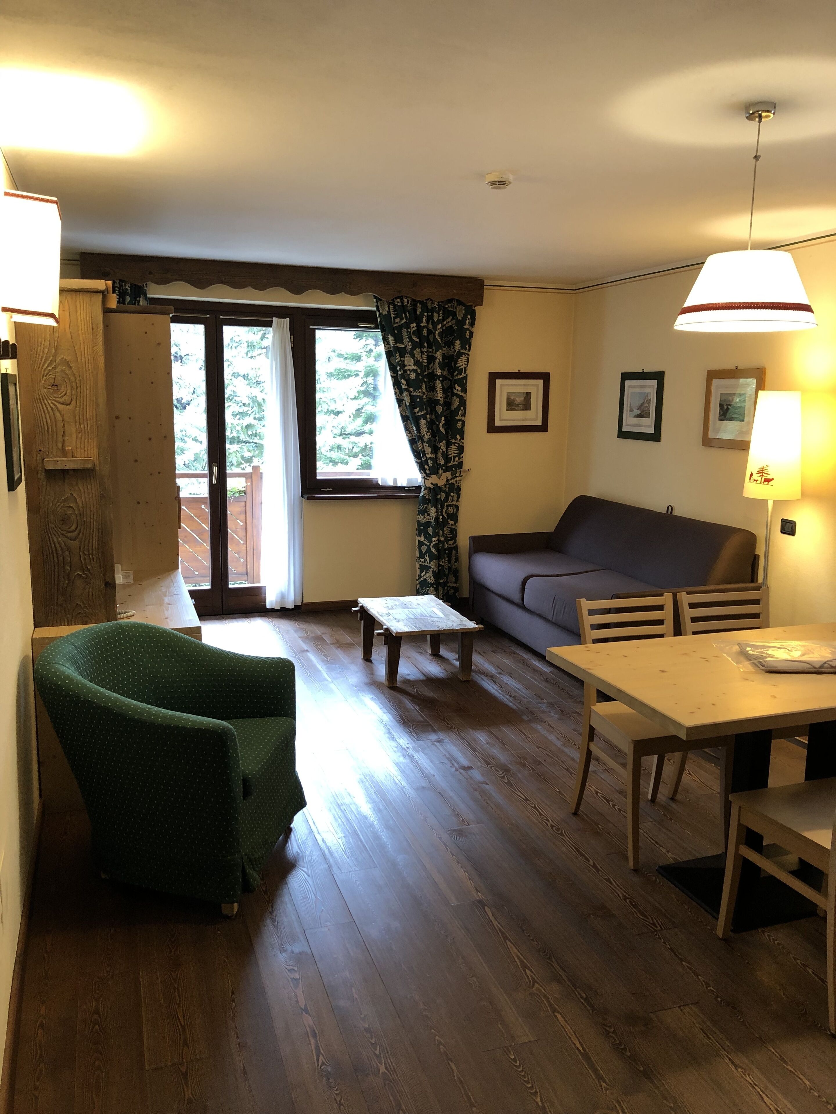 Ein Wohnzimmer mit Holzboden, einem grünen Sessel, einer Couch, einem kleinen Tisch und einer Lampe. Ein Fenster mit Vorhängen und eine Tür zu einem Außenbereich.