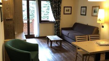 Ein Wohnzimmer mit Holzboden, einem grünen Sessel, einer Couch, einem kleinen Tisch und einer Lampe. Ein Fenster mit Vorhängen und eine Tür zu einem Außenbereich.