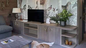 A modern living room with a flat-screen TV, wooden cabinets, a grey sofa, a coffee table, and a decorative wall with nature-themed wallpaper. - Residence Courmaison (Pre-Saint-Didier)