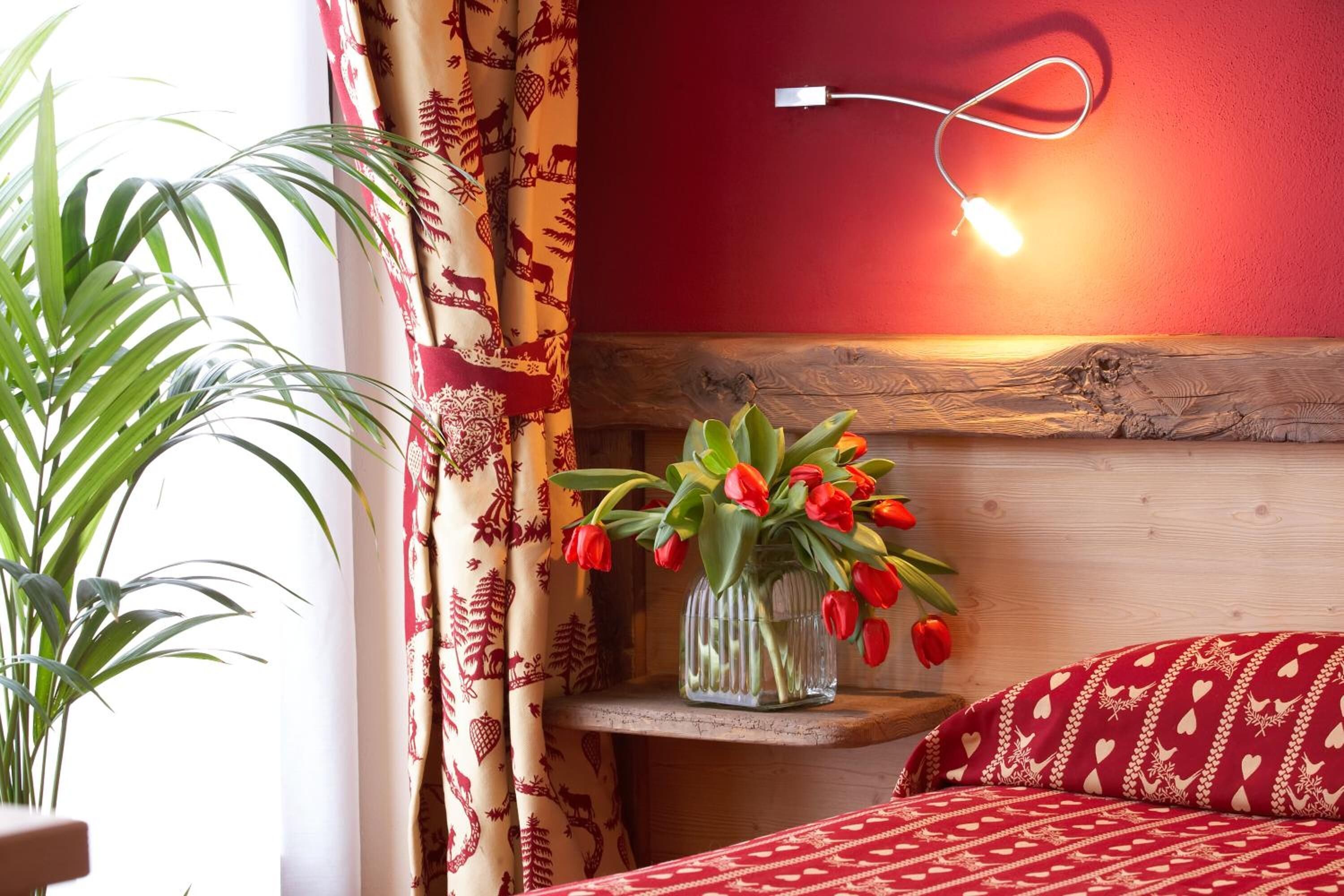 Una habitación con cabecera de madera, un florero con tulipanes rojos sobre una estantería, una colcha de cama con estampado rojo y una lámpara fijada a la pared.
