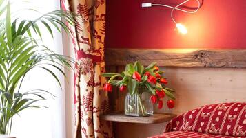 Una habitación con cabecera de madera, un florero con tulipanes rojos sobre una estantería, una colcha de cama con estampado rojo y una lámpara fijada a la pared.