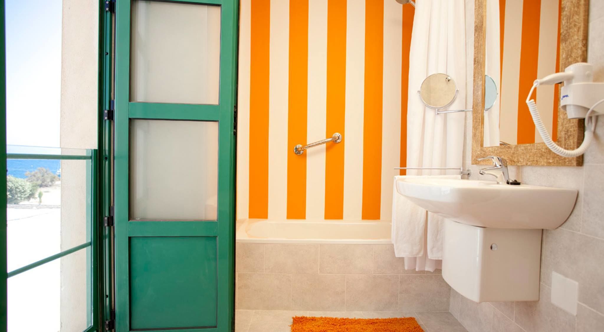 Un baño con puerta verde, pared rayada en naranja y blanco, lavamanos blanco y bañera.