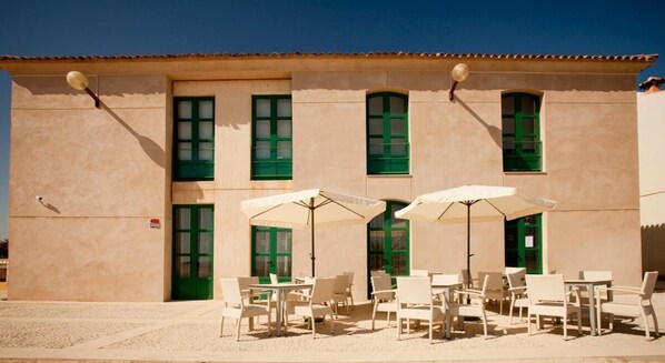 Un edificio con múltiples ventanas de marco verde y fachada color crema, que cuenta con asientos exteriores con sillas blancas y sombrillas.