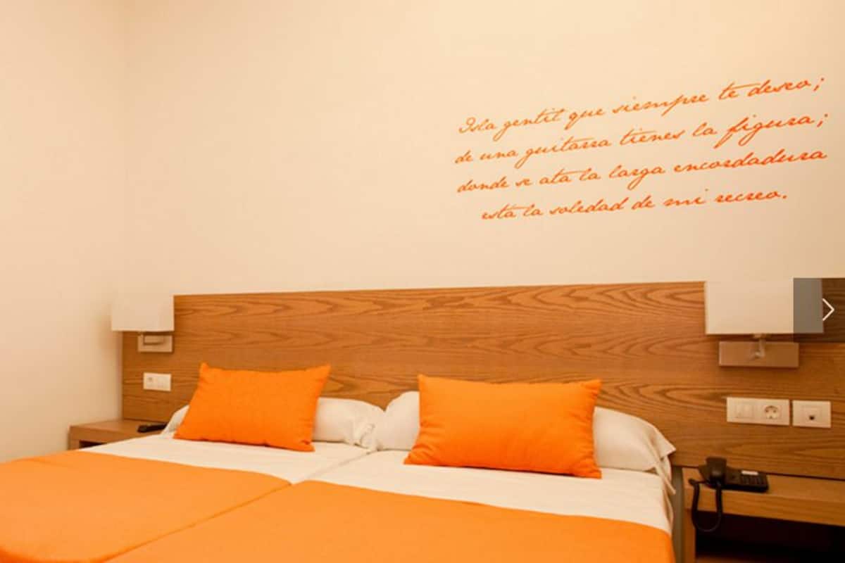 Habitación de hotel con cama, dos almohadas anaranjadas, cabecera de madera y vista a la playa.