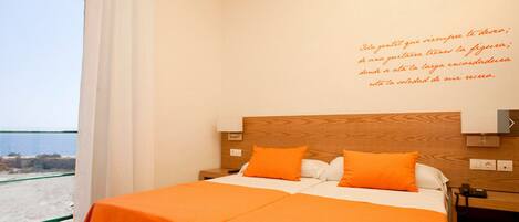 A hotel room with a bed, two orange pillows, a wooden headboard, and a view of the beach.