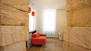 Une pièce aux murs de pierre, avec un canapé rouge, un fauteuil blanc et une grande fenêtre ornée de rideaux légers.
