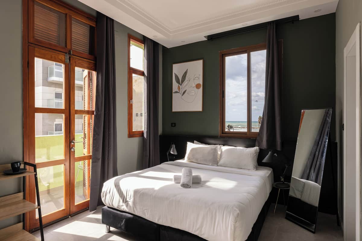 Un dormitorio con cama, ventana, un cuadro y vista a edificios.