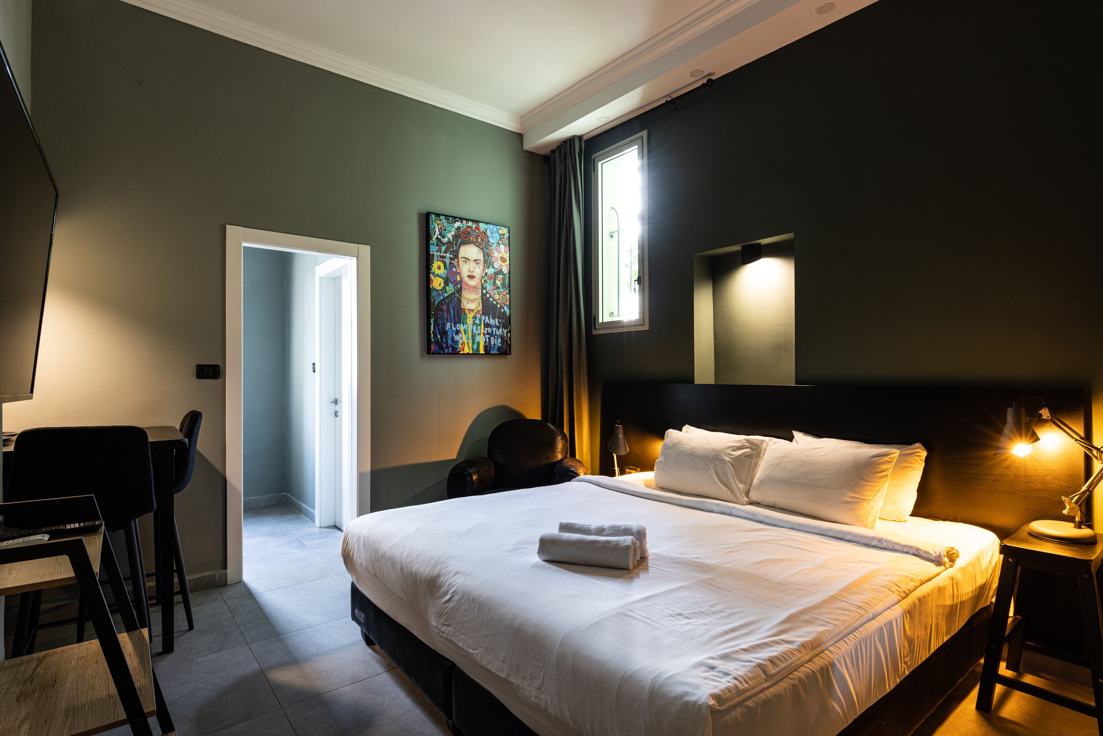 Ein Hotelzimmer mit einem ordentlich bezogenen Bett, einem Schreibtisch mit Stuhl, einem Bild an der Wand und einem Fenster mit Vorhängen.