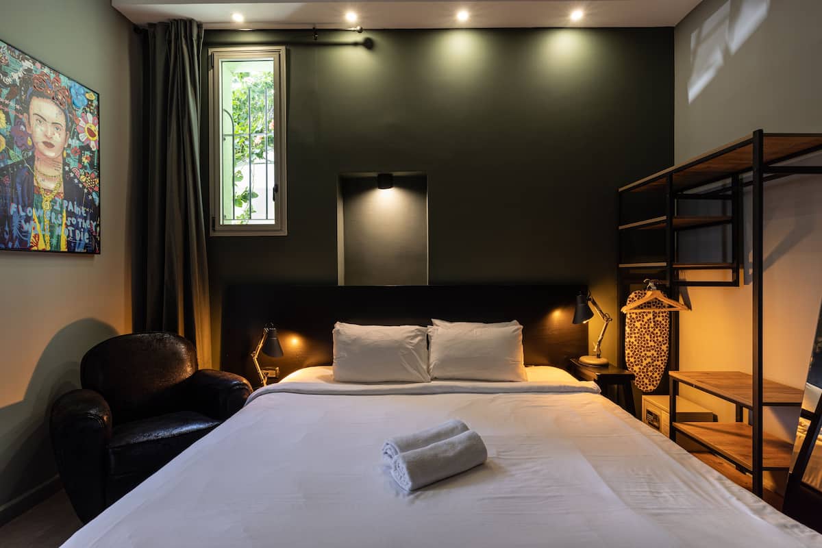 Una cama bien hecha con ropa blanca, una butaca de cuero negro, una mesita de noche con una lámpara y una ventana con cortinas.