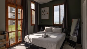 Un dormitorio con una cama grande, una ventana que ofrece vistas a edificios, un cuadro en la pared y una puerta que da a un balcón.