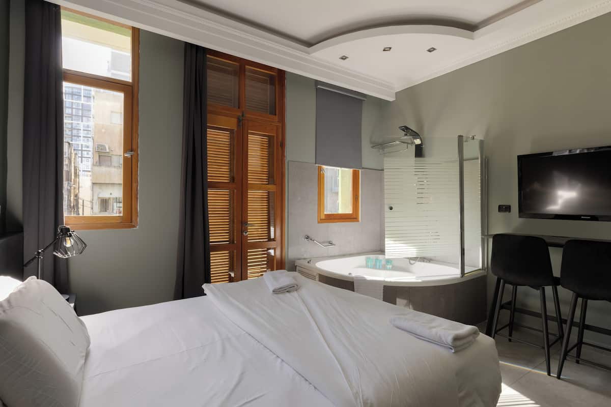 Una habitación de hotel con una cama grande, un baño con bañera, un televisor y dos sillones.