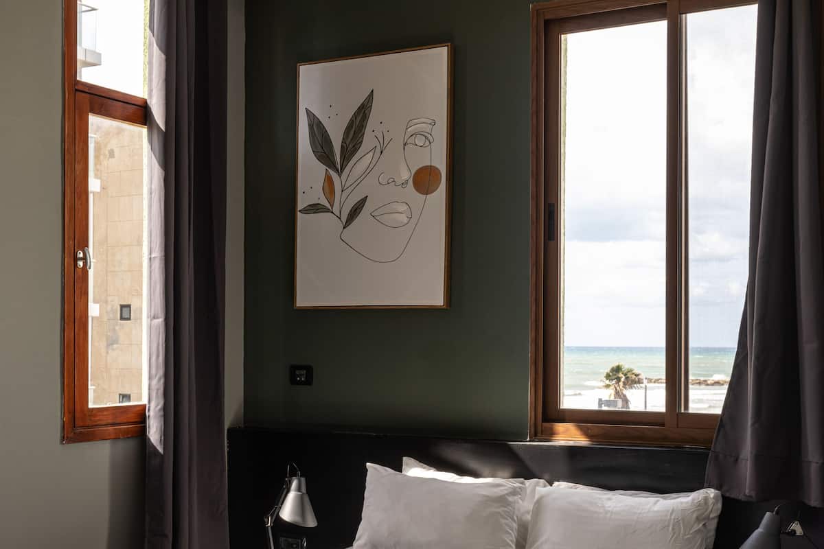 Una cama bien hecha con sábanas blancas, una obra de arte enmarcada en la pared y vista al mar por la ventana.
