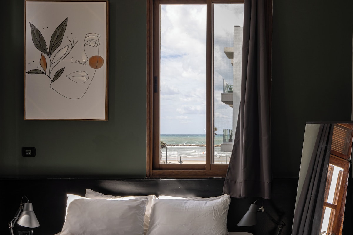 A hotel room with a bed, a window with a view of the beach, a framed artwork on the wall, and a mirror.