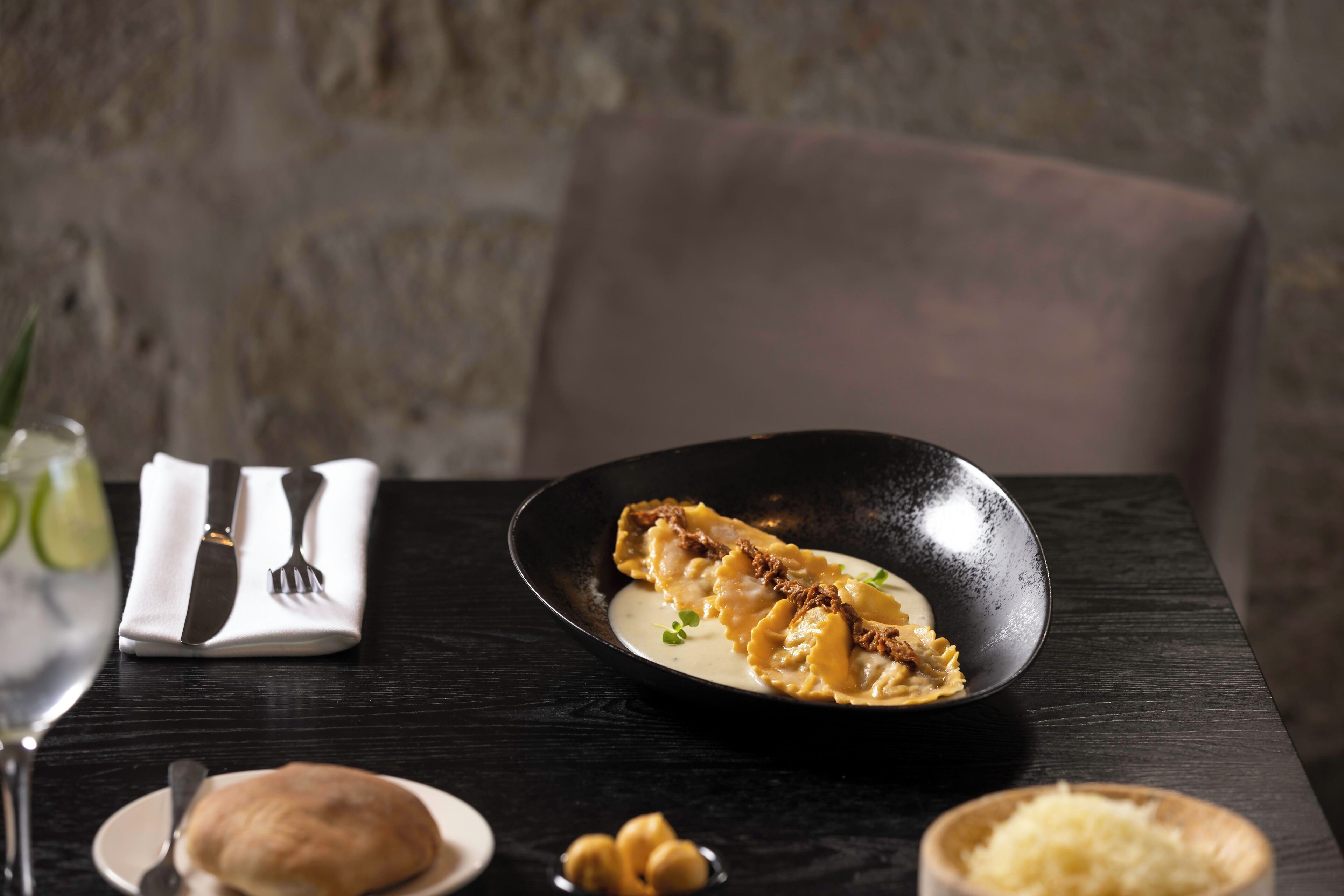 Una mesa con un plato de ravioles, un cuenco pequeño de queso rallado y una servilleta doblada con cubiertos.