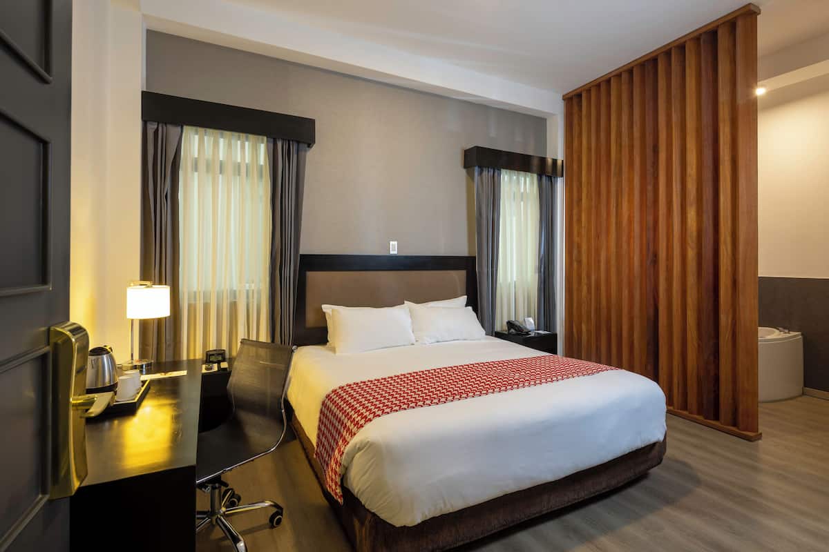 A hotel room with a large bed, a desk with a lamp, a chair, a nightstand, and a wooden partition.