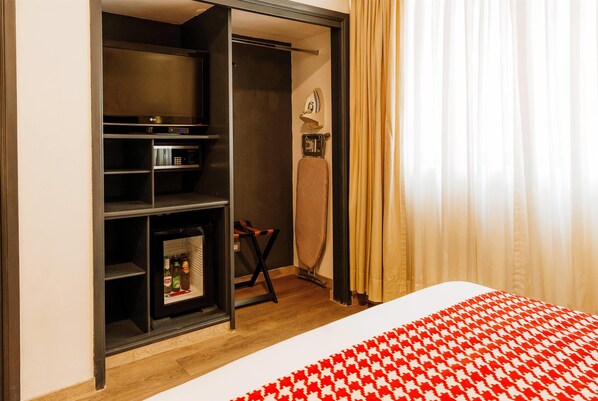 Una habitación de hotel con armario, cama con colcha de cuadros rojo y blanco, y un pequeño rincón de estar con una silla y una mesa.