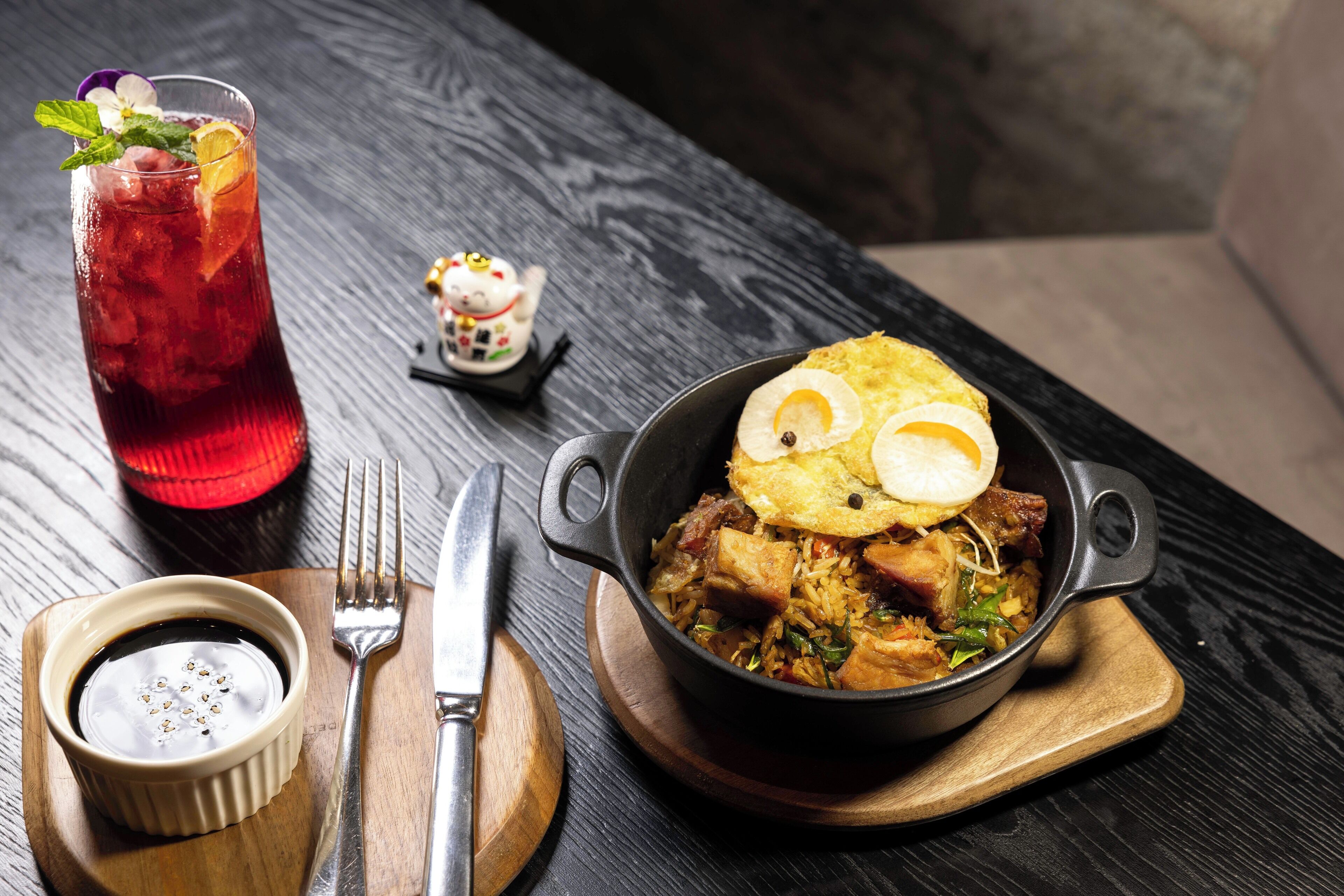 Un repas composé de riz, de viande et d’un œuf au plat, servi sur une planche en bois, accompagné d’un bol de soupe et d’un verre de thé glacé, le tout posé sur une table.