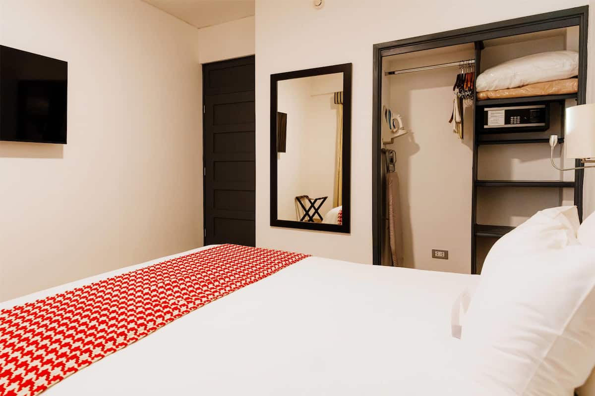 A hotel room with a bed, a red and white patterned bedspread, a mirror, a wardrobe, and a shower area.