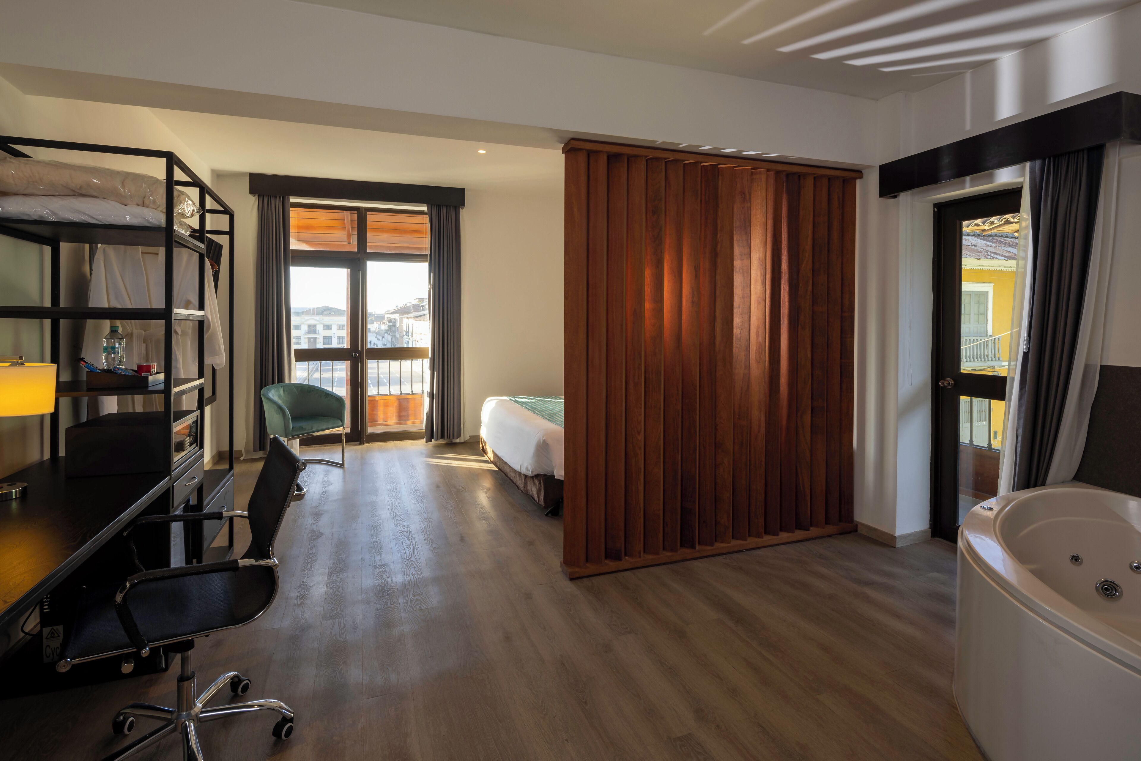 A hotel room with a wooden floor, a desk with a chair, a bunk bed, a large window, a sliding door, and a bathtub.