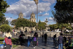 Una plaza pública con una fuente, edificios históricos y gente caminando.