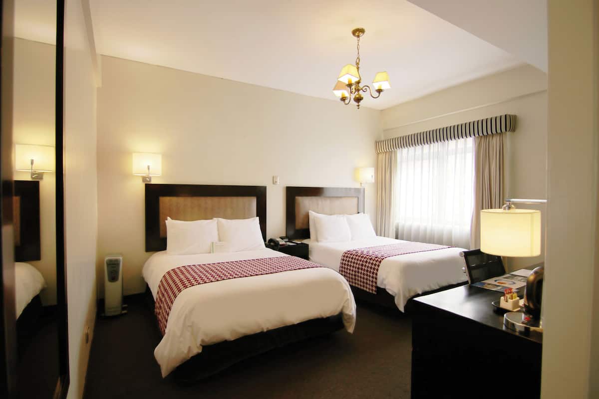 A hotel room with two beds, a desk, a chair, a lamp, and a window with curtains.