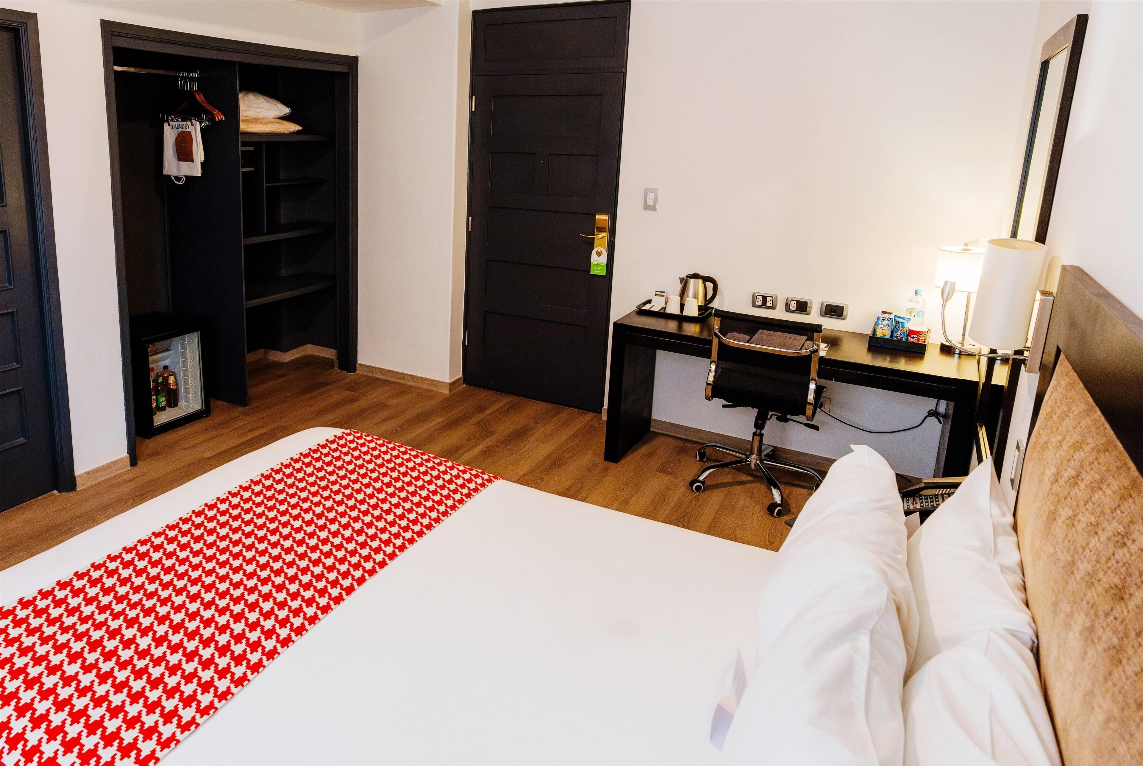 Habitación de hotel con cama, escritorio, silla, armario y mesita de noche con lámpara.