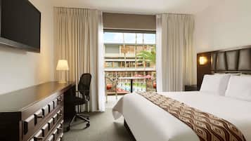 Habitación de hotel con cama, escritorio, silla, televisor y balcón con vista a un edificio y palmeras.