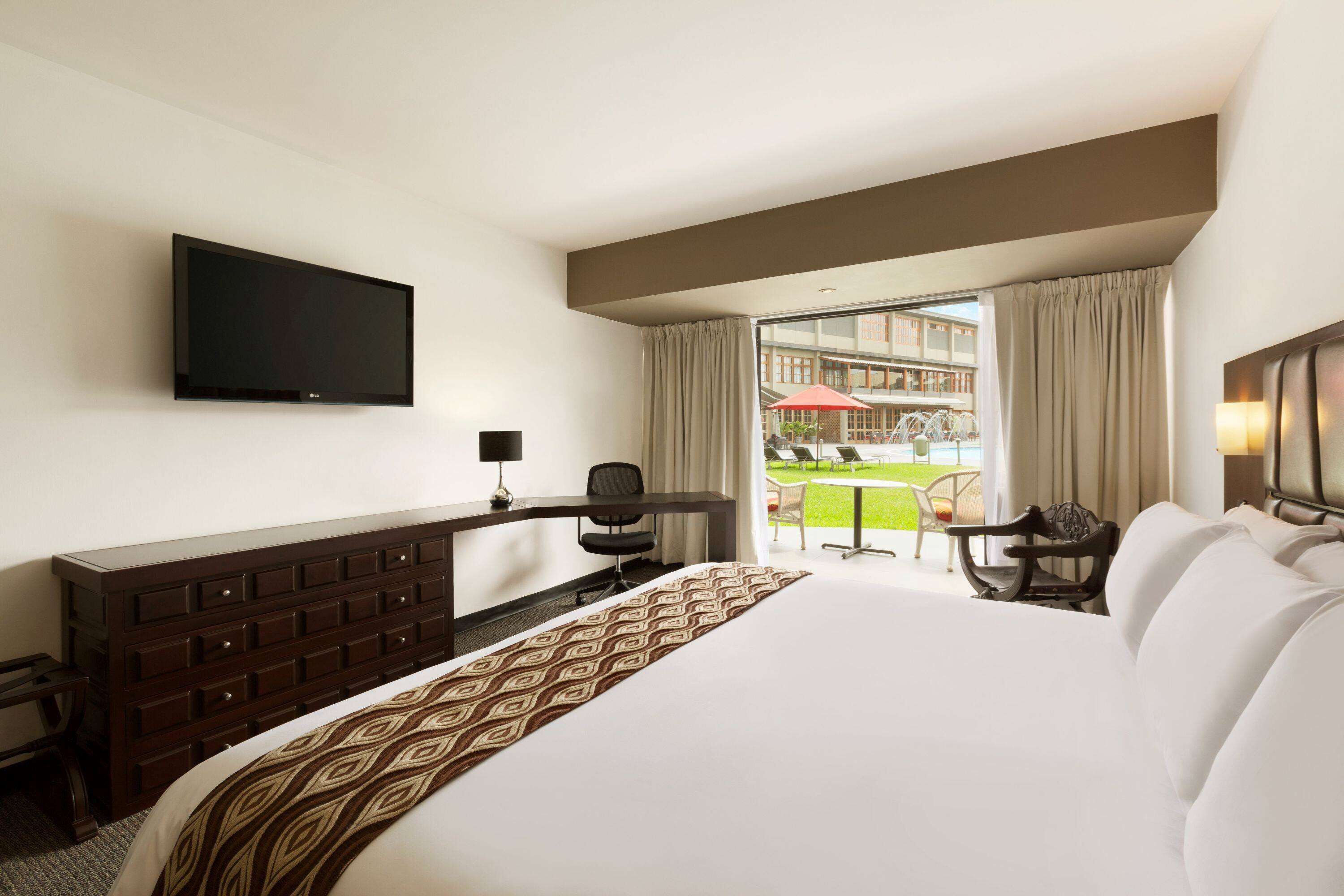 Habitación de hotel con una cama grande, un televisor montado en la pared, un escritorio con una silla y vistas a un espacio exterior con mesa y sillas.