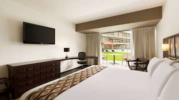 Habitación de hotel con una cama grande, un televisor montado en la pared, un escritorio con una silla y vistas a un espacio exterior con mesa y sillas.