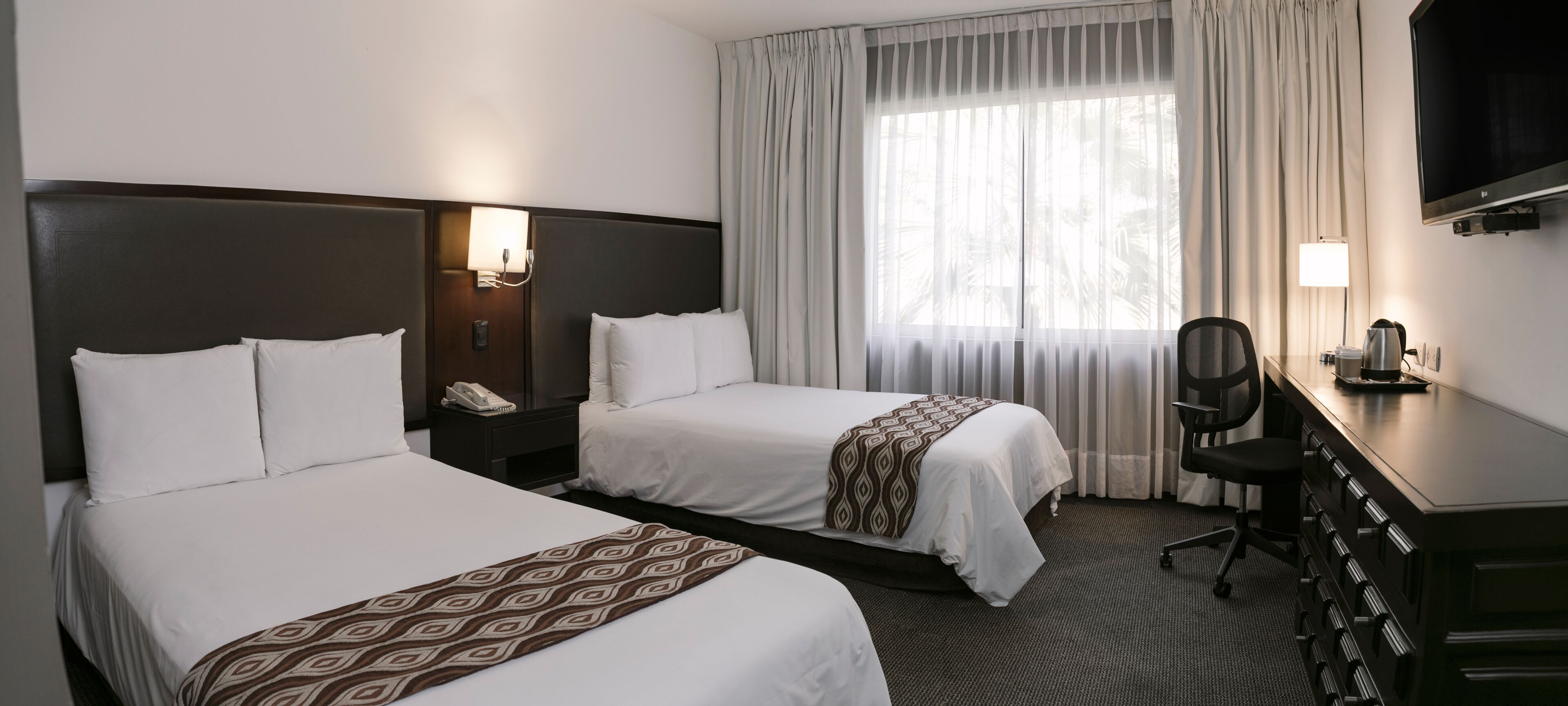 Habitación de hotel con dos camas, un escritorio con silla, televisor y ventana con cortinas.