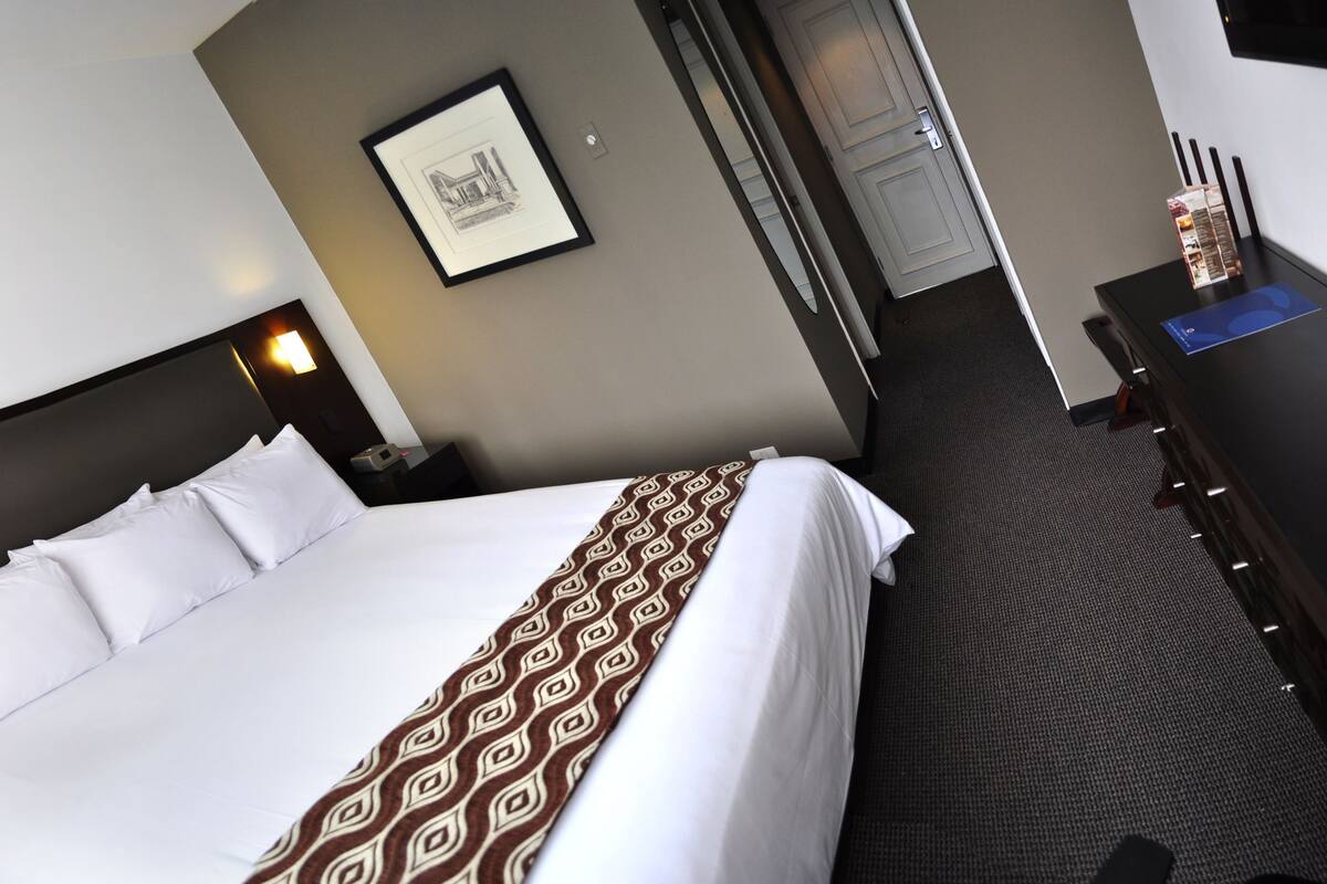 A hotel room with a large bed, a bedside table with a phone, a framed picture on the wall, and a desk with a computer and a box of tissues.