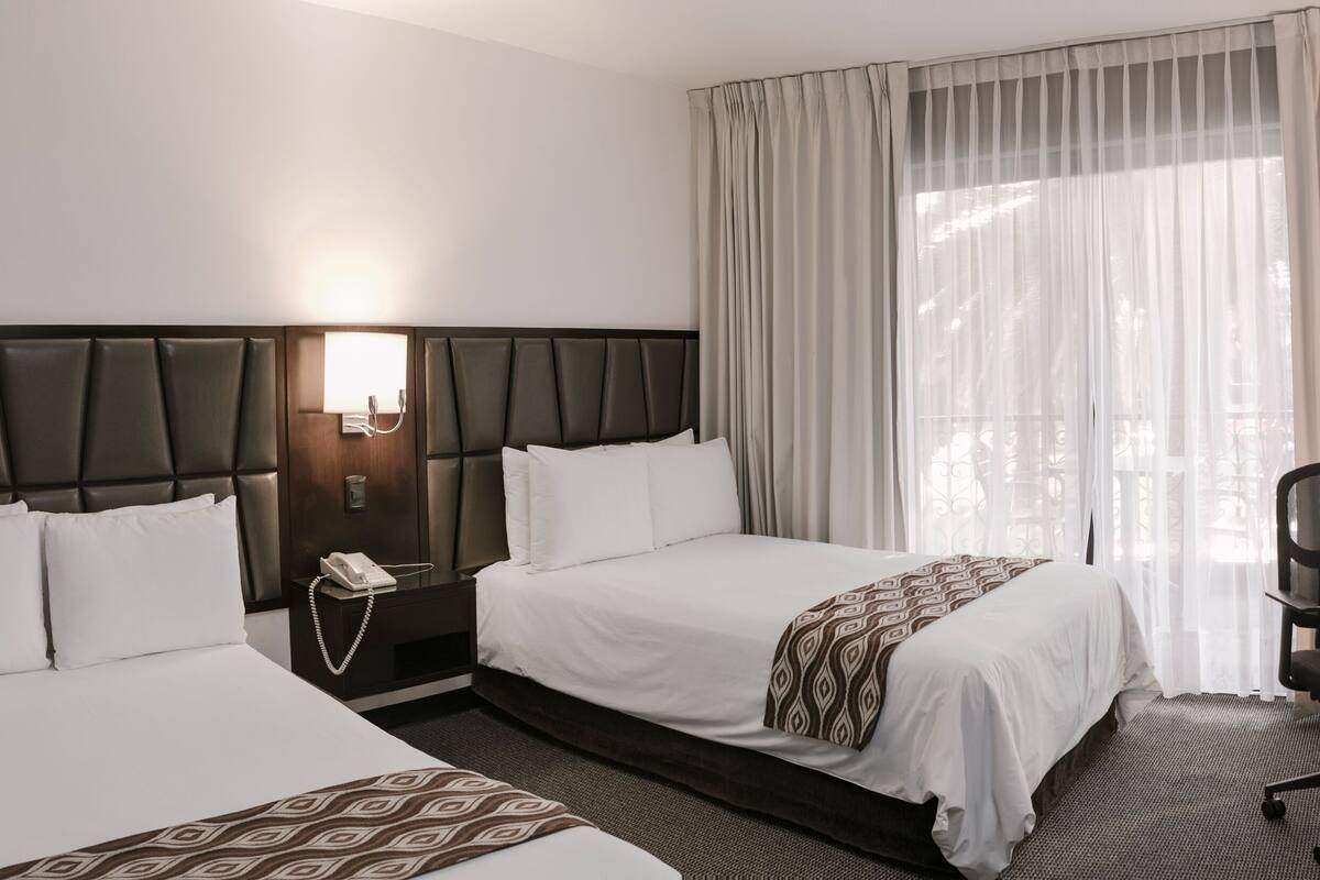 A hotel room with two beds, a desk, a chair, a TV, and a window with curtains.