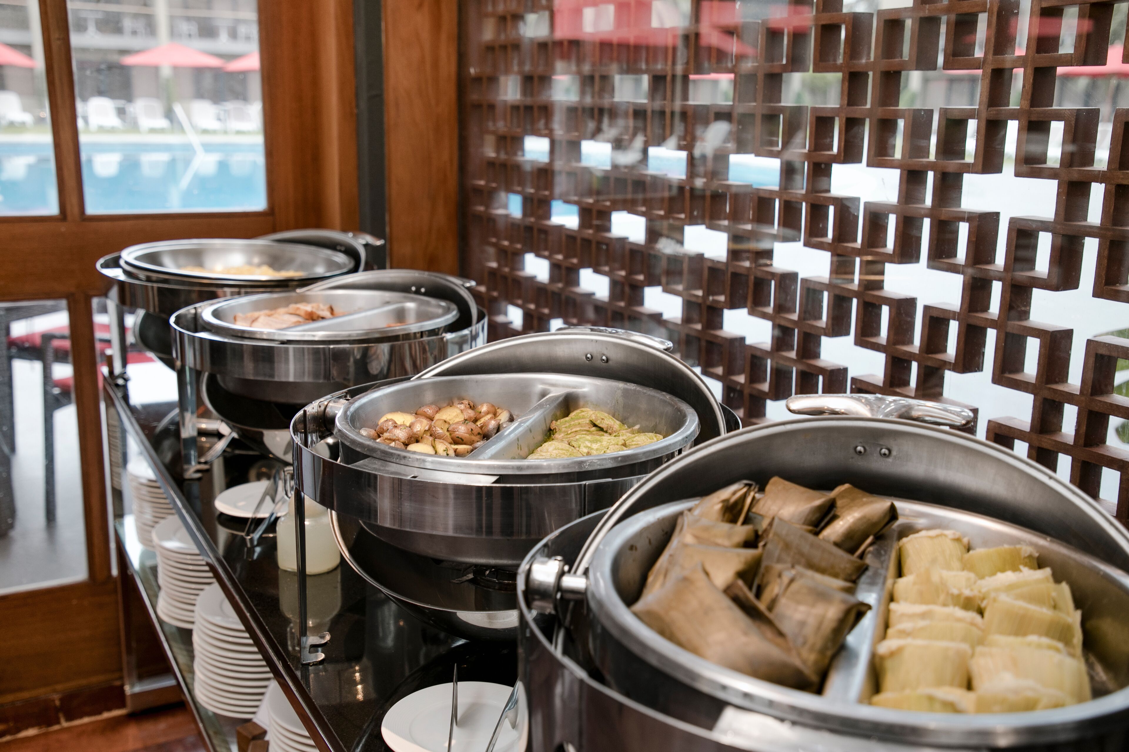 A buffet with various food items in metal containers, a glass display case with plates, and a view of the outside through a window with a wooden lattice.