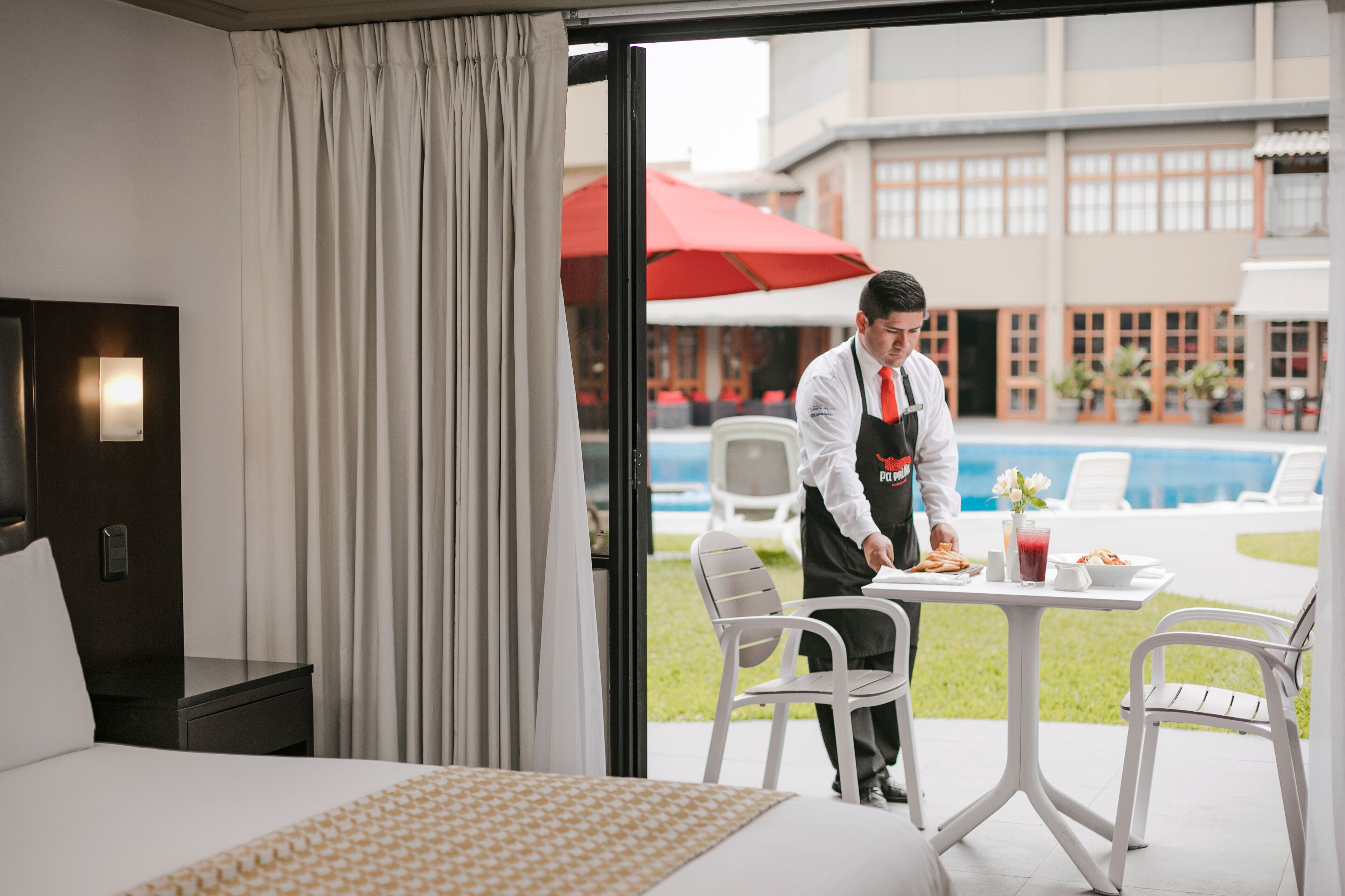 A hotel room with a bed, a dining table with chairs, a red umbrella, and a pool in the background.