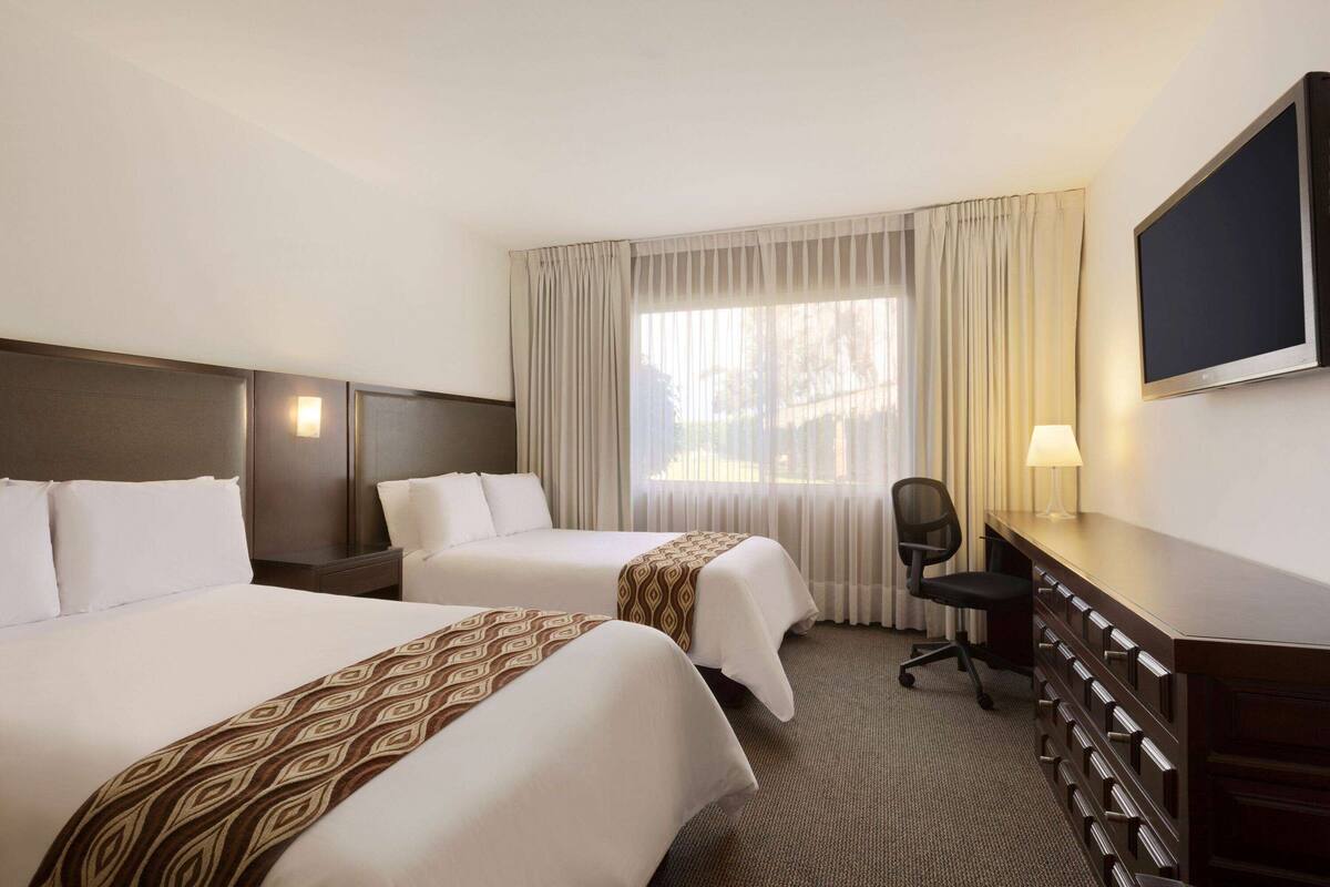 A hotel room with two beds, a desk, a chair, a TV, and a window with curtains.