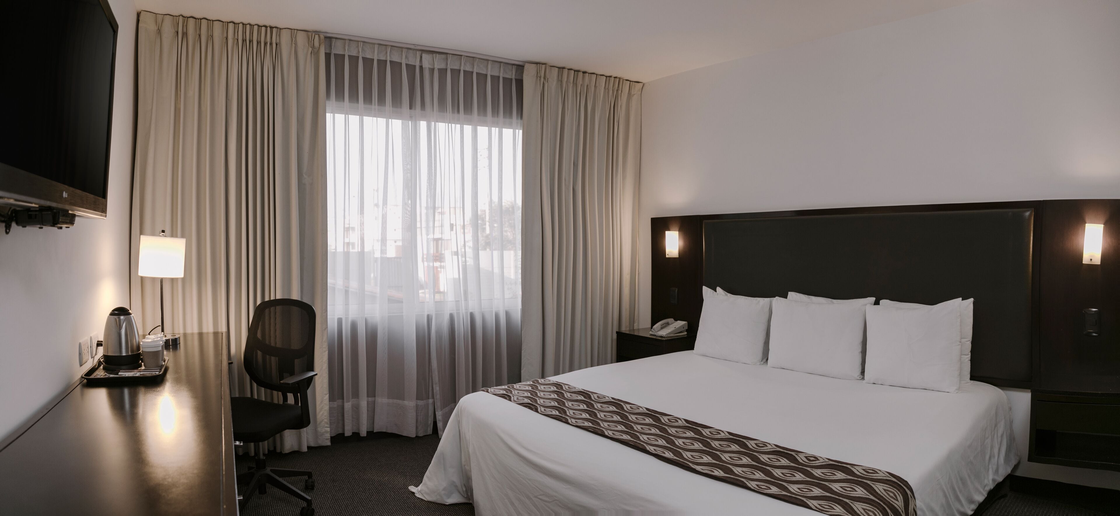 Habitación de hotel con una cama grande, un escritorio con cafetera, una silla, televisión y ventana con cortinas.