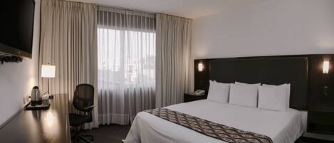 Habitación de hotel con una cama grande, un escritorio con cafetera, una silla, televisión y ventana con cortinas.