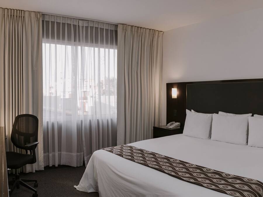 A hotel room with a large bed, a desk with a coffee maker, a chair, a TV, and a window with curtains.