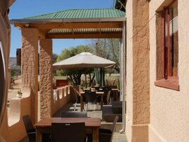 Un patio exterior con pilares de piedra, mesas y sillas de madera, y un paraguas grande.