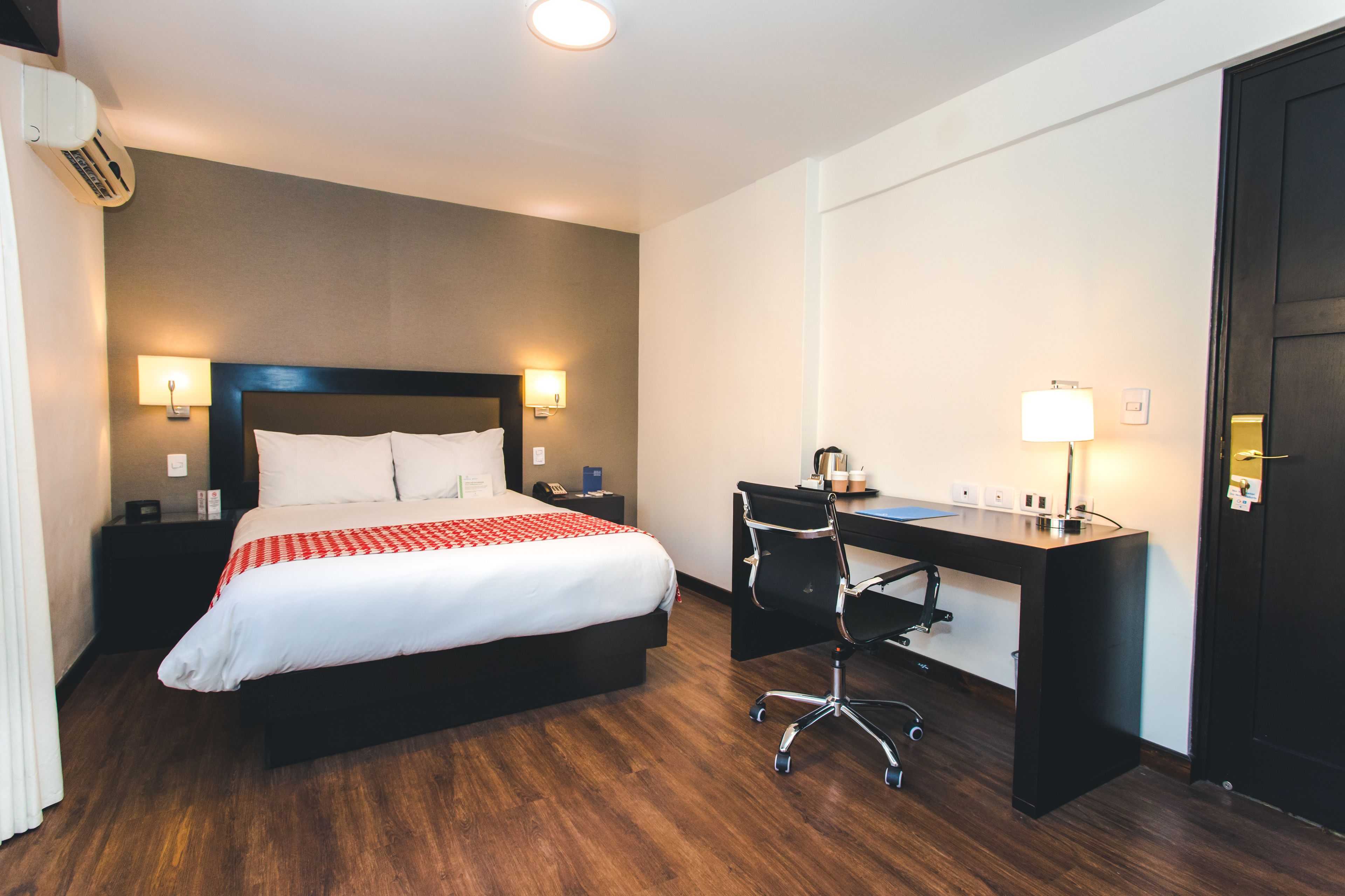A hotel room with a large bed, a desk, and a chair. There are two wall-mounted lamps and a desk lamp.