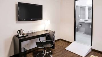A hotel room with a flat-screen TV, a desk with a chair, a bedside lamp, and a view of the bathroom through a glass door.