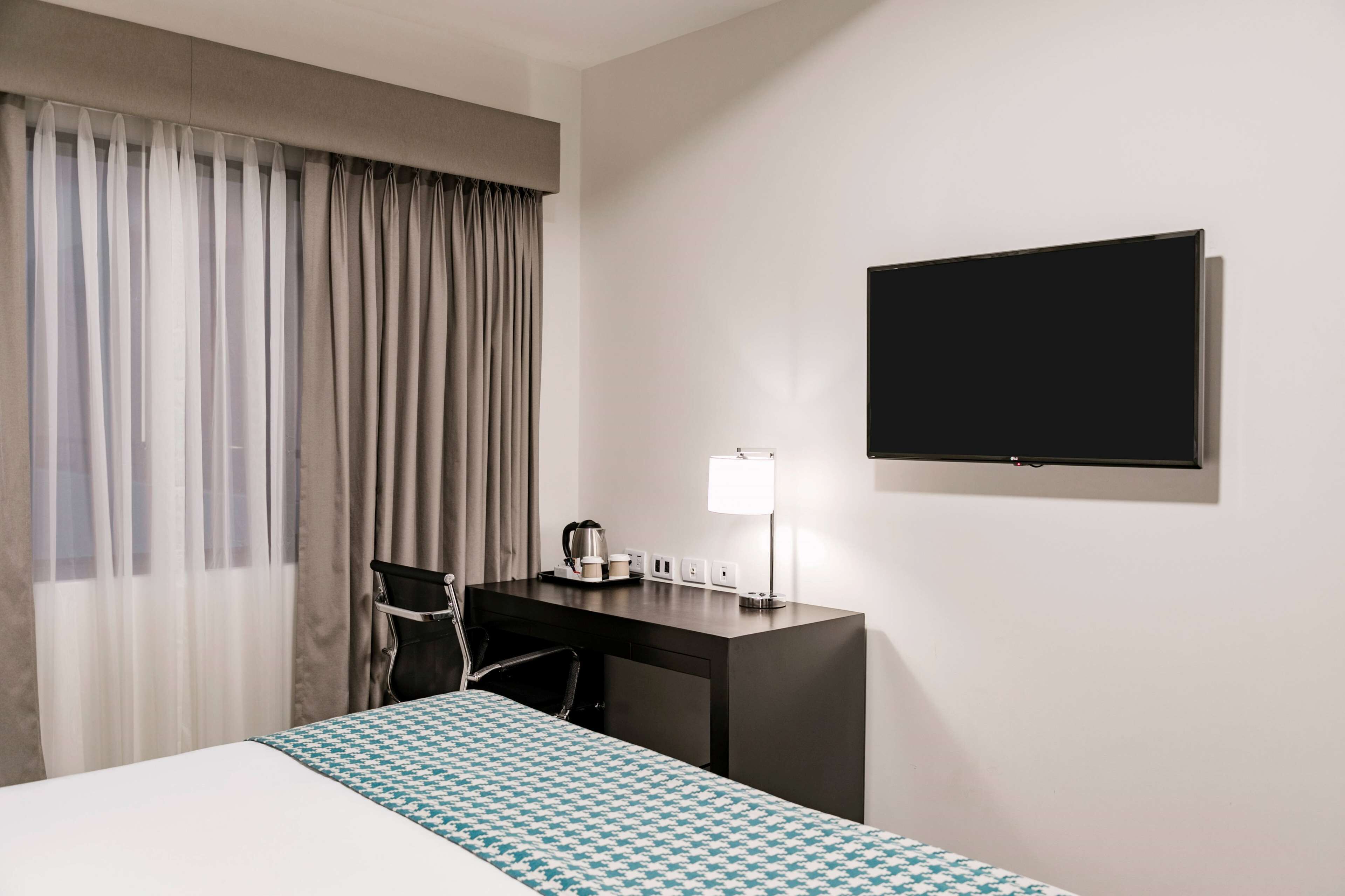 Ett hotellrum med en säng, ett skrivbord med en lampa, en stol, en tv och ett fönster med gardiner.
