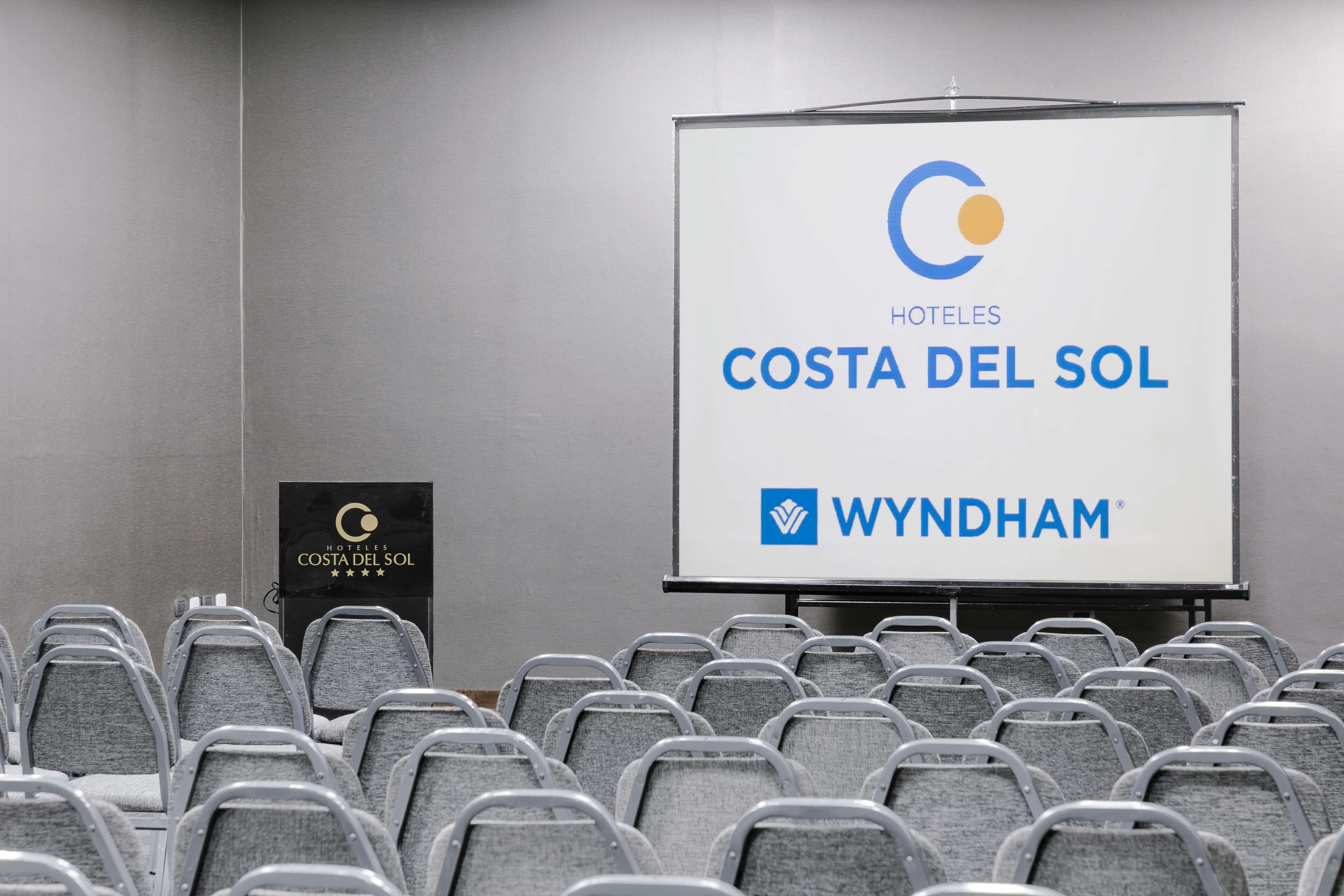 Un salón de conferencias con filas de sillas grises, una pantalla de proyección mostrando los logotipos de 'HOTELES COSTA DEL SOL' y 'WYNDHAM', y un atril con el mismo nombre de hotel.